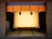 A traditional Japanese room with tatami mat flooring and a wooden frame at the center. The frame is adorned with intricate golden designs at the corners. Above it is a decorative hanging with a blue and orange pattern featuring tassels in black and red.