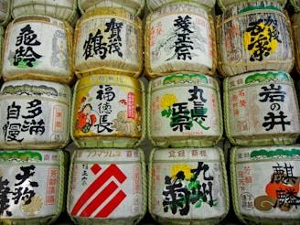 A collection of traditional Japanese sake barrels adorned with calligraphy and intricate designs, arranged in neat rows. Each barrel is wrapped in straw and features bold, colorful text and imagery, depicting cultural symbols and characters.