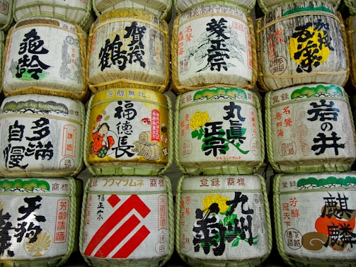 A collection of traditional Japanese sake barrels adorned with calligraphy and intricate designs, arranged in neat rows. Each barrel is wrapped in straw and features bold, colorful text and imagery, depicting cultural symbols and characters.