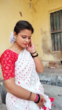 A woman is standing, wearing a traditional white sari with red patterns and a red blouse. She has ornaments like earrings and a necklace, with flowers in her hair. The background shows an old, slightly worn wall with a window.