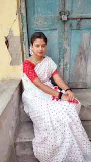 A model wearing a traditional white and red Bengali saree, standing by a rustic wooden door.