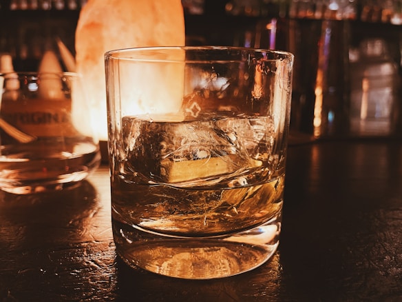 A clear glass filled with whiskey and large ice cubes sits on a dimly lit bar counter. Warm lighting creates a cozy ambiance, reflecting off the glass surface. There is a subtle glow from a light source in the background, suggesting a relaxed, intimate setting.