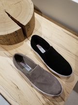 A pair of casual slip-on shoes is placed on a wooden surface. One shoe is black and the other is gray. They have a simple, minimalist design with labels visible on the inner sole. Nearby is a wooden object with a natural, rustic texture and visible grain.