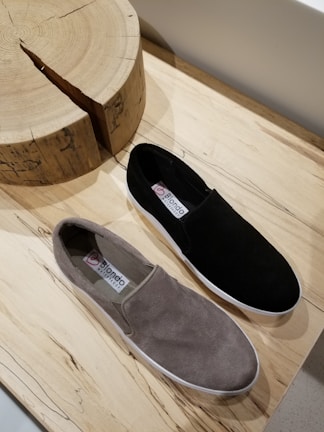 A pair of casual slip-on shoes is placed on a wooden surface. One shoe is black and the other is gray. They have a simple, minimalist design with labels visible on the inner sole. Nearby is a wooden object with a natural, rustic texture and visible grain.
