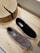 A pair of casual slip-on shoes is placed on a wooden surface. One shoe is black and the other is gray. They have a simple, minimalist design with labels visible on the inner sole. Nearby is a wooden object with a natural, rustic texture and visible grain.