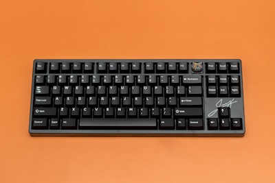 A black mechanical keyboard with white lettering is set against an orange background. The keyboard features a compact layout, including a custom keycap with a design in the top right corner and a signature on the bottom right.