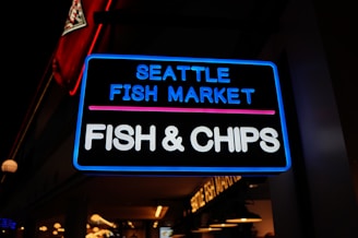 A neon sign brightly illuminated at night, advertising the Seattle Fish Market with a focus on Fish & Chips. The background shows a dimly lit interior with hanging lights, and part of a red, white, and black logo is visible in the corner.