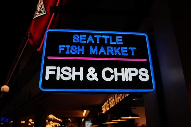A neon sign brightly illuminated at night, advertising the Seattle Fish Market with a focus on Fish & Chips. The background shows a dimly lit interior with hanging lights, and part of a red, white, and black logo is visible in the corner.