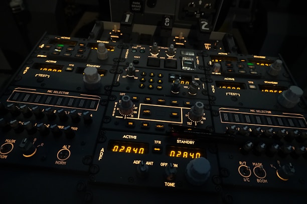 A complex aircraft cockpit control panel with multiple dials, switches, and illuminated displays. The panel features labels such as 'ACTIVE', 'STANDBY', and 'MIC SELECTOR', with various numerical readouts and settings. The ambient lighting is dim, highlighting the glowing indicators and knobs.
