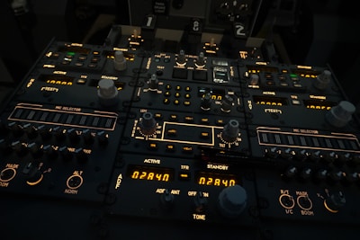A complex aircraft cockpit control panel with multiple dials, switches, and illuminated displays. The panel features labels such as 'ACTIVE', 'STANDBY', and 'MIC SELECTOR', with various numerical readouts and settings. The ambient lighting is dim, highlighting the glowing indicators and knobs.