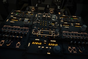 A complex aircraft cockpit control panel with multiple dials, switches, and illuminated displays. The panel features labels such as 'ACTIVE', 'STANDBY', and 'MIC SELECTOR', with various numerical readouts and settings. The ambient lighting is dim, highlighting the glowing indicators and knobs.