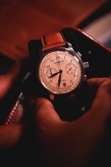 A detailed wristwatch with a leather strap is held by a pair of hands. The watch face is cream-colored with black numerals and features additional dials for seconds and minutes. The lighting casts a warm glow, emphasizing the craftsmanship and elegance of the timepiece.