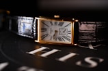 A rectangular wristwatch with a white dial, featuring Roman numerals and a small subdial for seconds. The watch has a gold case and a black leather strap. It is placed on a surface with the word 'HIS' visible in the background.