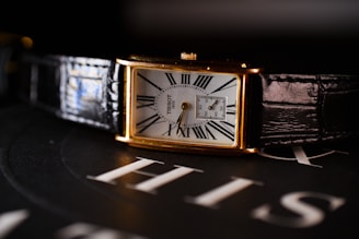 A rectangular wristwatch with a white dial, featuring Roman numerals and a small subdial for seconds. The watch has a gold case and a black leather strap. It is placed on a surface with the word 'HIS' visible in the background.