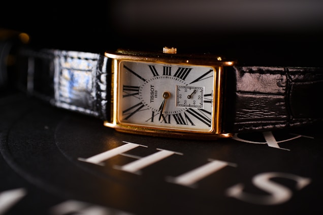 A rectangular wristwatch with a white dial, featuring Roman numerals and a small subdial for seconds. The watch has a gold case and a black leather strap. It is placed on a surface with the word 'HIS' visible in the background.