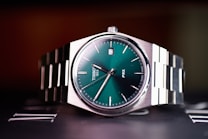 A close-up view of a stainless steel wristwatch with a green dial and silver metallic bracelet. The watch face displays minimalistic hour markers and a date window at the 3 o'clock position. The brand name and watch model are visible on the dial.