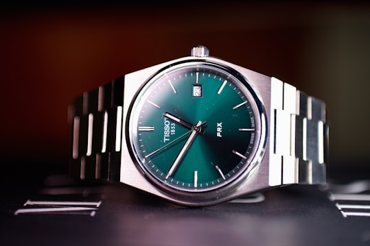 A close-up view of a stainless steel wristwatch with a green dial and silver metallic bracelet. The watch face displays minimalistic hour markers and a date window at the 3 o'clock position. The brand name and watch model are visible on the dial.