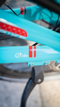 Close-up view of a turquoise electric bike frame with red stripes and the word 'bike' along with an electrical plug symbol. The bike is supported by a black stand, and parts of the wheel are visible.