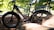 A black e-bike is parked outdoors, partially obscured by rocks and surrounded by a lush green forest. Sunlight filters through the leaves, casting dappled shadows on the ground and bike. The bike features thick tires and a sturdy frame, suitable for off-road use.