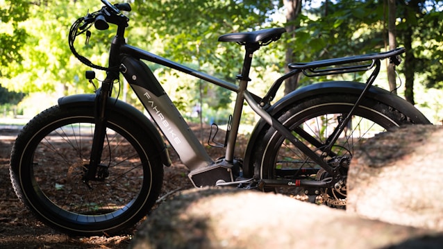 A black e-bike is parked outdoors, partially obscured by rocks and surrounded by a lush green forest. Sunlight filters through the leaves, casting dappled shadows on the ground and bike. The bike features thick tires and a sturdy frame, suitable for off-road use.