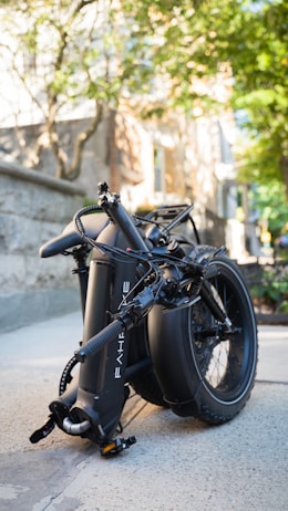 A black bicycle, likely an electric bike, seen folded and placed on a sidewalk. The background includes a stone wall, trees, and some buildings, suggesting an urban setting with a mix of natural and constructed elements.