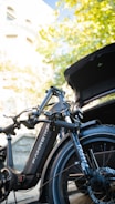 A sleek black electric bike is positioned near an open car trunk, partially under the cover of vibrant green leaves and against a backdrop of a sunny building exterior.