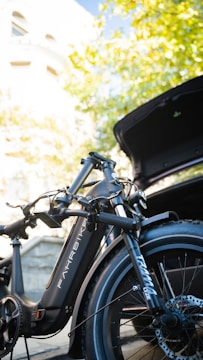 A sleek black electric bike is positioned near an open car trunk, partially under the cover of vibrant green leaves and against a backdrop of a sunny building exterior.