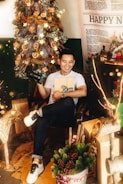 A cheerful person is sitting in a festive room, holding a camera and a wrapped gift. The background features a decorated Christmas tree with lights and ornaments. Various wrapped presents are placed around, along with some festive decorations such as pinecones and branches. There is a large 'Happy New' poster in the backdrop, partially visible, suggesting a holiday or Christmas setting.