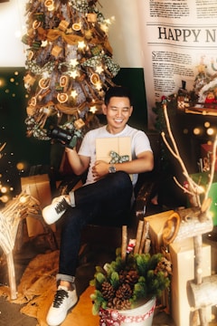 A cheerful person is sitting in a festive room, holding a camera and a wrapped gift. The background features a decorated Christmas tree with lights and ornaments. Various wrapped presents are placed around, along with some festive decorations such as pinecones and branches. There is a large 'Happy New' poster in the backdrop, partially visible, suggesting a holiday or Christmas setting.
