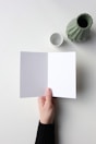 A hand holding an open card with one blank white page and one blank gray page, placed on a light surface. Nearby, a green vase and a small white cup are positioned.