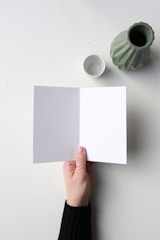 A hand holding an open card with one blank white page and one blank gray page, placed on a light surface. Nearby, a green vase and a small white cup are positioned.
