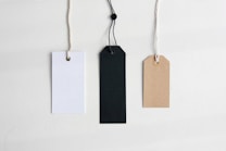 Three blank tags with strings are aligned side by side on a neutral background. The tags are rectangular in shape and vary in color, including white, black, and brown.