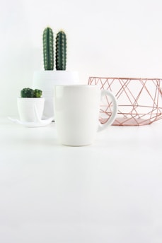 A minimalist scene featuring a white ceramic mug in the foreground, flanked by potted cacti in white planters. In the background, a decorative geometric wire basket adds a touch of modern style.