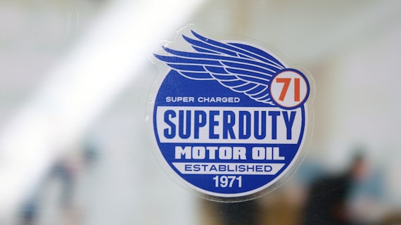 A sticker with a blue circular design features the text 'Superduty Motor Oil' along with the phrases 'Super Charged' and 'Established 1971.' A stylized wing and the number '71' are incorporated into the design.