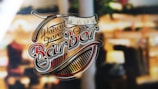 A vintage-style logo for a barbershop featuring ornate lettering and images of a comb and straight razor against a blurred background, suggesting a shop window.