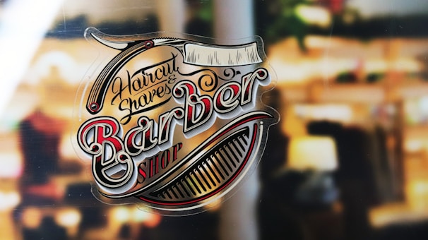 A vintage-style logo for a barbershop featuring ornate lettering and images of a comb and straight razor against a blurred background, suggesting a shop window.