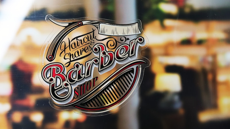 A vintage-style logo for a barbershop featuring ornate lettering and images of a comb and straight razor against a blurred background, suggesting a shop window.