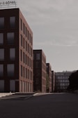A series of modern, rectangular buildings with a uniform architecture line a quiet, empty street. The buildings are mostly composed of red brick with large glass windows, and the overcast sky adds a muted tone to the scene. A sign with the word 'BÖRSE' is visible at the top of one of the buildings.