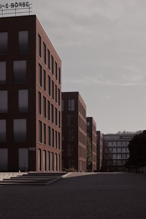A series of modern, rectangular buildings with a uniform architecture line a quiet, empty street. The buildings are mostly composed of red brick with large glass windows, and the overcast sky adds a muted tone to the scene. A sign with the word 'BÖRSE' is visible at the top of one of the buildings.