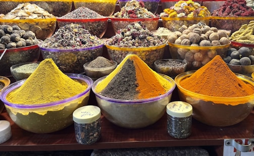 Various colorful spices are showcased in large, clear bowls arranged neatly on a market stall. The spices include vibrant yellow, orange, and brown hues, surrounded by an assortment of dried herbs, fruits, and flowers. Smaller jars are placed in front, containing more spices or herbs. The setup suggests a bustling marketplace with an array of aromatic and visually appealing ingredients.