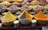 Various colorful spices are showcased in large, clear bowls arranged neatly on a market stall. The spices include vibrant yellow, orange, and brown hues, surrounded by an assortment of dried herbs, fruits, and flowers. Smaller jars are placed in front, containing more spices or herbs. The setup suggests a bustling marketplace with an array of aromatic and visually appealing ingredients.