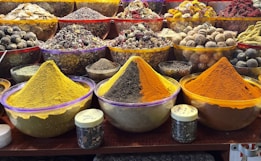 Various colorful spices are showcased in large, clear bowls arranged neatly on a market stall. The spices include vibrant yellow, orange, and brown hues, surrounded by an assortment of dried herbs, fruits, and flowers. Smaller jars are placed in front, containing more spices or herbs. The setup suggests a bustling marketplace with an array of aromatic and visually appealing ingredients.
