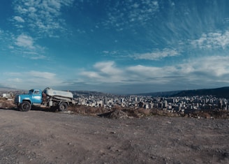 A vintage blue truck with a tanker is parked on a rocky, dirt terrain overlooking a sprawling cityscape. The city stretches across the valley under a vast blue sky dotted with wispy clouds.