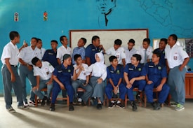 A group of young people, likely students, are gathered indoors in front of a blue decorated wall featuring a map and a portrait. They are wearing uniforms with some in white shirts and others in blue. A mix of standing and seated poses, they appear to be interacting and smiling at each other, suggesting a casual, friendly atmosphere.