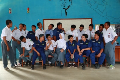 A group of young people, likely students, are gathered indoors in front of a blue decorated wall featuring a map and a portrait. They are wearing uniforms with some in white shirts and others in blue. A mix of standing and seated poses, they appear to be interacting and smiling at each other, suggesting a casual, friendly atmosphere.