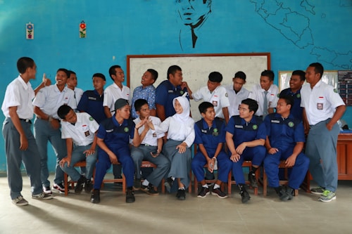 A group of young people, likely students, are gathered indoors in front of a blue decorated wall featuring a map and a portrait. They are wearing uniforms with some in white shirts and others in blue. A mix of standing and seated poses, they appear to be interacting and smiling at each other, suggesting a casual, friendly atmosphere.