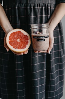 A person holds a halved grapefruit in one hand and a jar labeled 'Joy Grapefruit Scrub' in the other. The individual is wearing a dark dress with a checkered pattern in light pink.