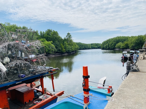 Fishing boats are docked along a calm river bordered by lush green mangroves under a partly cloudy sky. A heap of fishing nets and gear is visible on the left, while the waterway stretches into the distance.