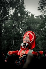 A performer is dressed in a vibrant traditional costume, characterized by a large, ornate headpiece and red, black, and white face paint. The costume is elaborate with intricate patterns, including circular designs on the headpiece. The background is a natural setting with lush green trees, suggesting an outdoor event or festival.
