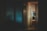 A dimly lit office environment with an open door leading into a room. There are papers hanging on the wall, a desk with a stack of files, and a partially visible computer screen.